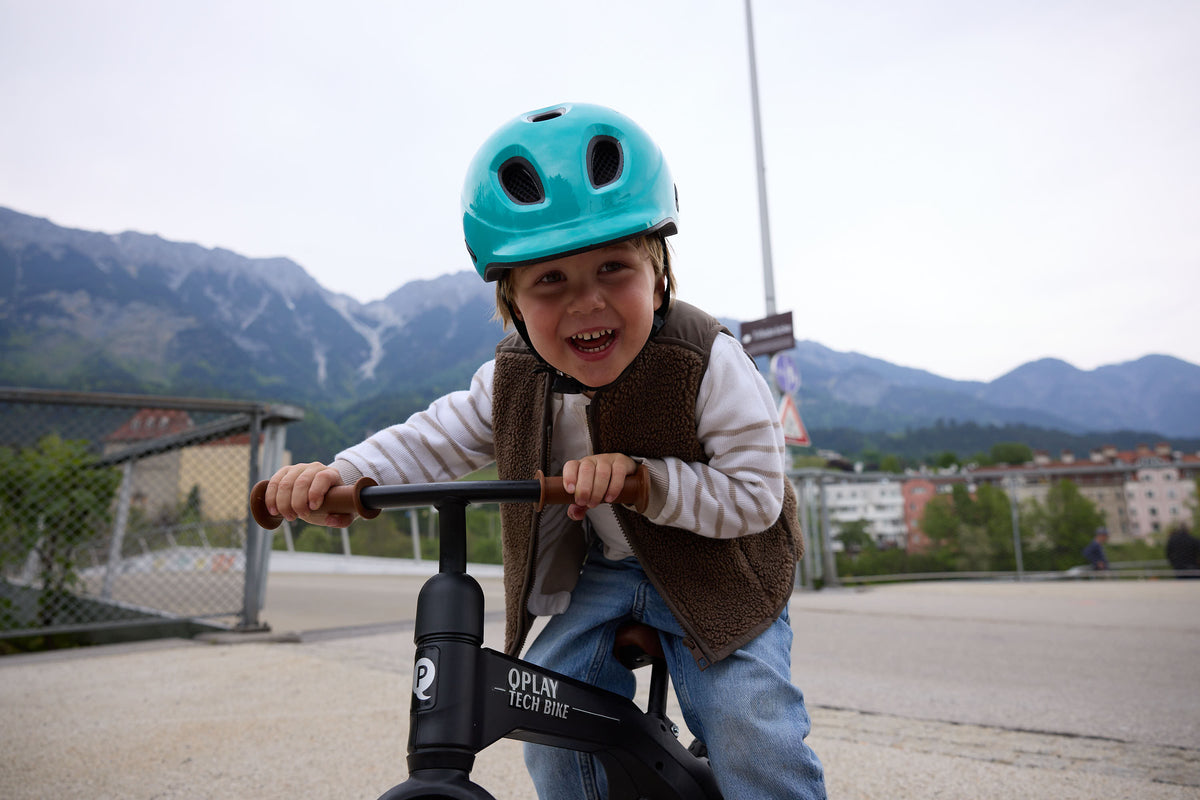 Kind auf Laufrad mit türkisem KED Kinderhelm vor Bergkulisse